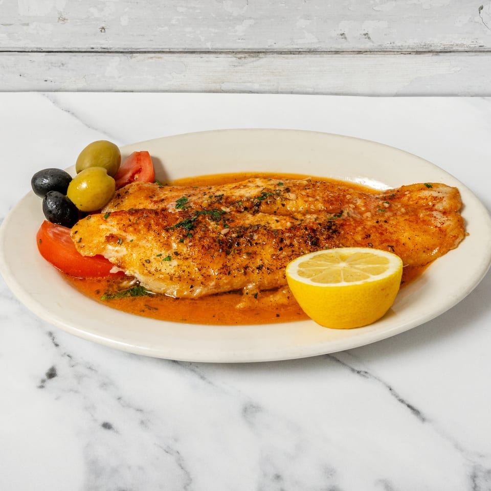 Seasoned fish fillet in orange sauce with olives, tomato slices, and a lemon wedge.