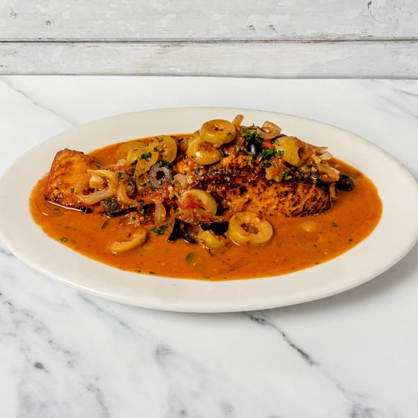 Seared fish in a rich tomato and olive sauce with onions and herbs on a white plate.