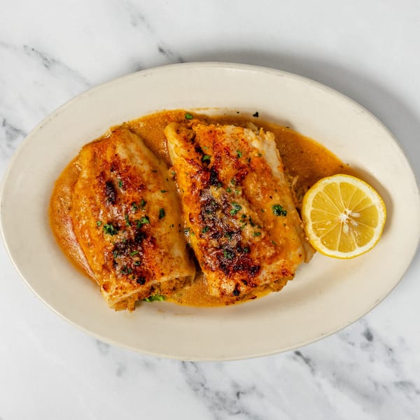 Two baked fish fillets in a rich orange sauce with parsley and a lemon slice on a white plate.