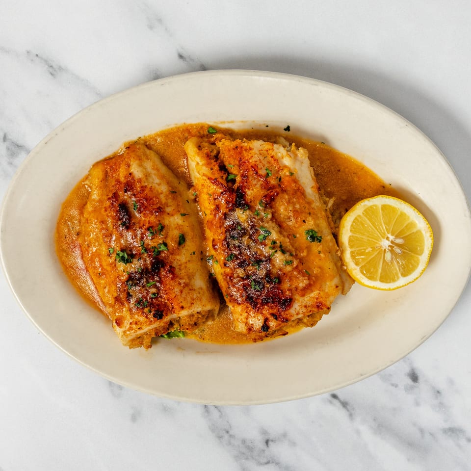 Two baked fish fillets in a rich orange sauce with parsley and a lemon slice on a white plate.