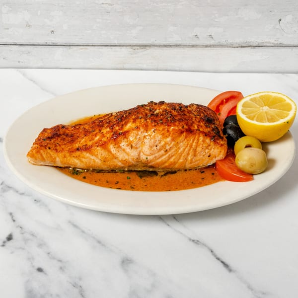 Grilled salmon fillet with orange sauce, lemon, tomatoes, and olives on a white plate.
