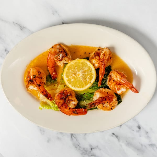 Plate of grilled shrimp, lemon, and greens in a buttery sauce on a marble surface.