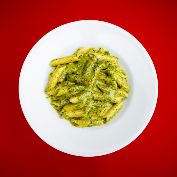 Penne pasta coated in creamy green pesto sauce on a white plate against a red background.