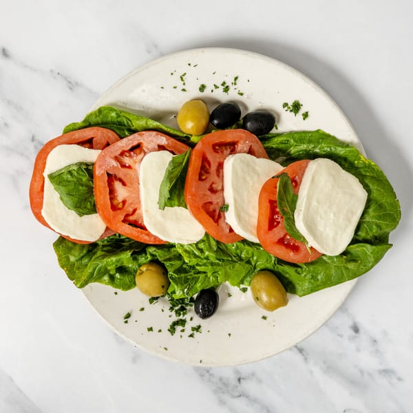 Caprese salad with sliced tomatoes, mozzarella, basil, lettuce, and olives on a white plate.