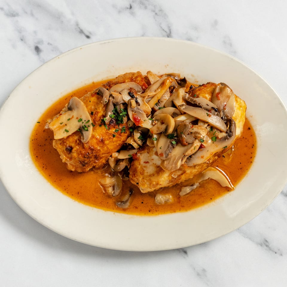 Two golden-brown chicken breasts topped with sliced mushrooms and a rich reddish-orange sauce on a white plate.